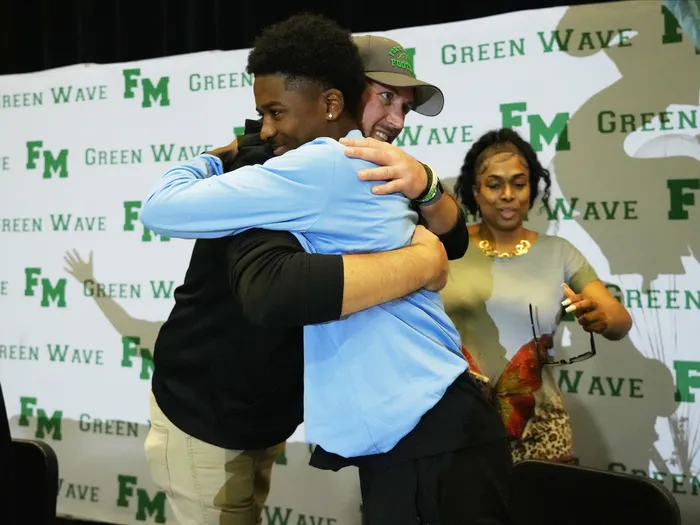 Emotional moment during a high school signing event as a student-athlete in a blue shirt hugs his coach on stage. A woman smiles beside them, and a branded 'Green Wave FM' backdrop is behind them.