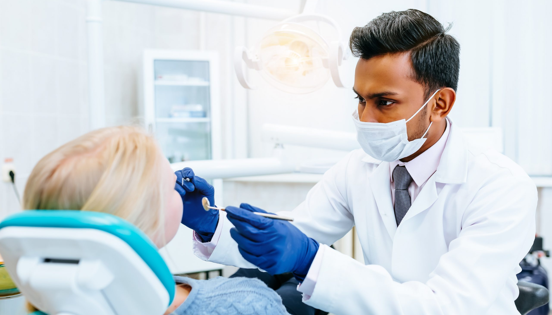 Tannspesialist på Eiksmarka – Når trenger du én? 16 Tannlege med munnbind og hansker undersøker pasientens tenner i en moderne klinikk, pasienten sitter tilbakelent i blå behandlingsstol.