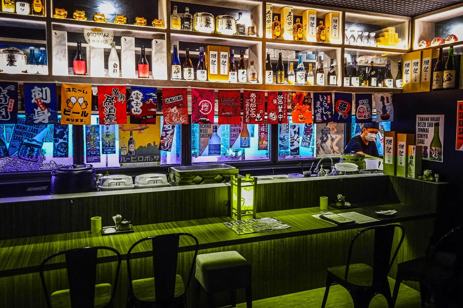 Cozy Japanese bar interior with wooden counters, colorful banners, and shelves of sake bottles. Warm lighting creates an inviting atmosphere.