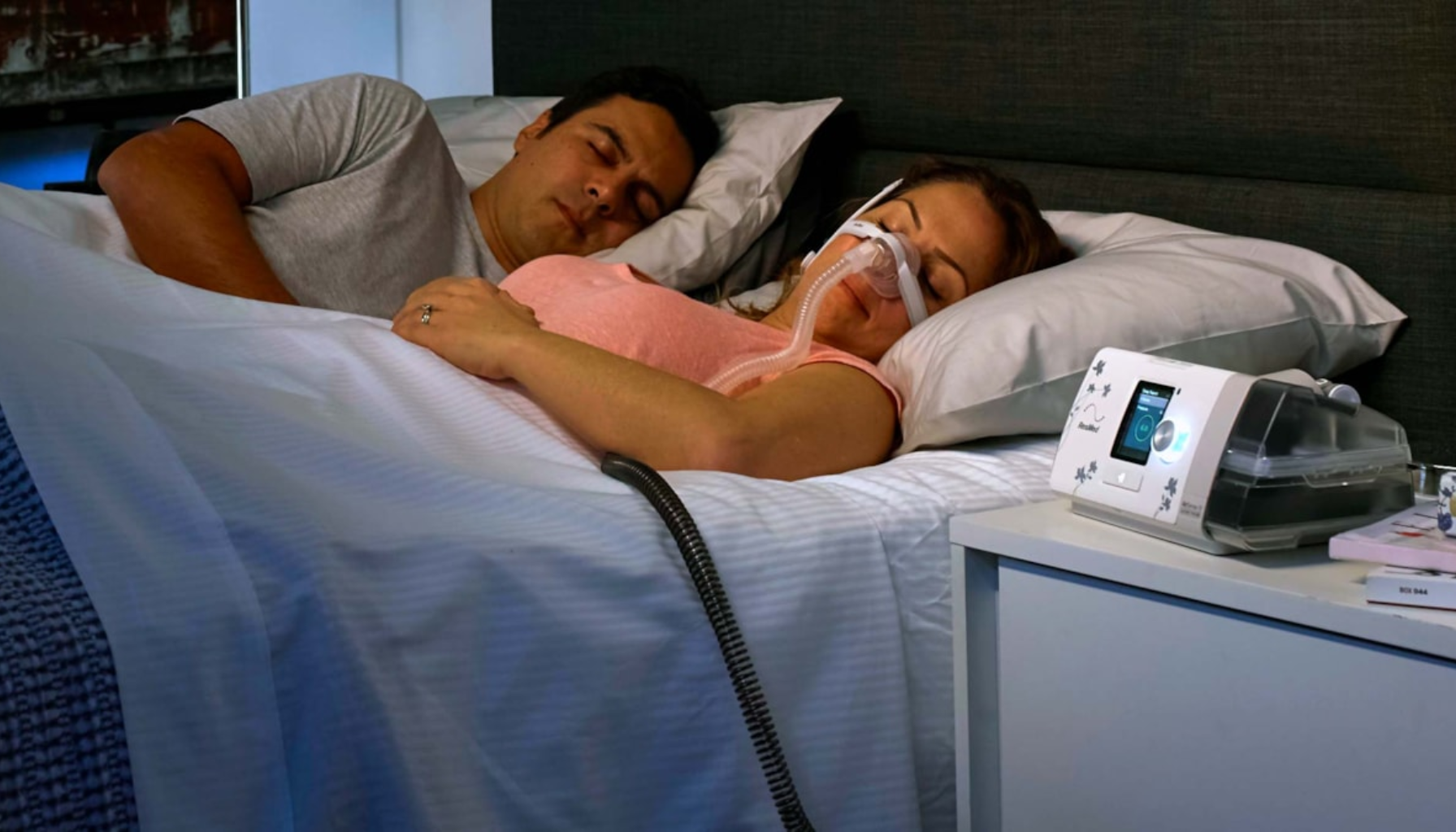 Kvinne sover med CPAP-maskin mot søvnapné mens partner ligger ved siden av i sengen.