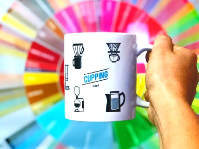 Pessoa segura caneca com desenhos de cafeteiras e fundo colorido da Cupping Café. Foto: Reprodução/instagram.com/cuppingcafe/