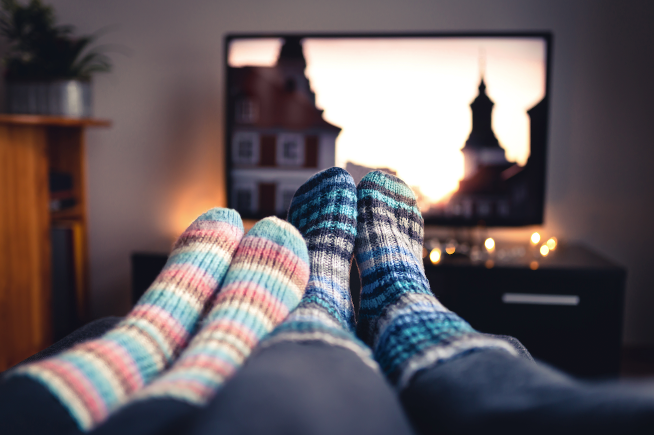 two pairs of feet in cozy socks are propped up with a television playing in the background