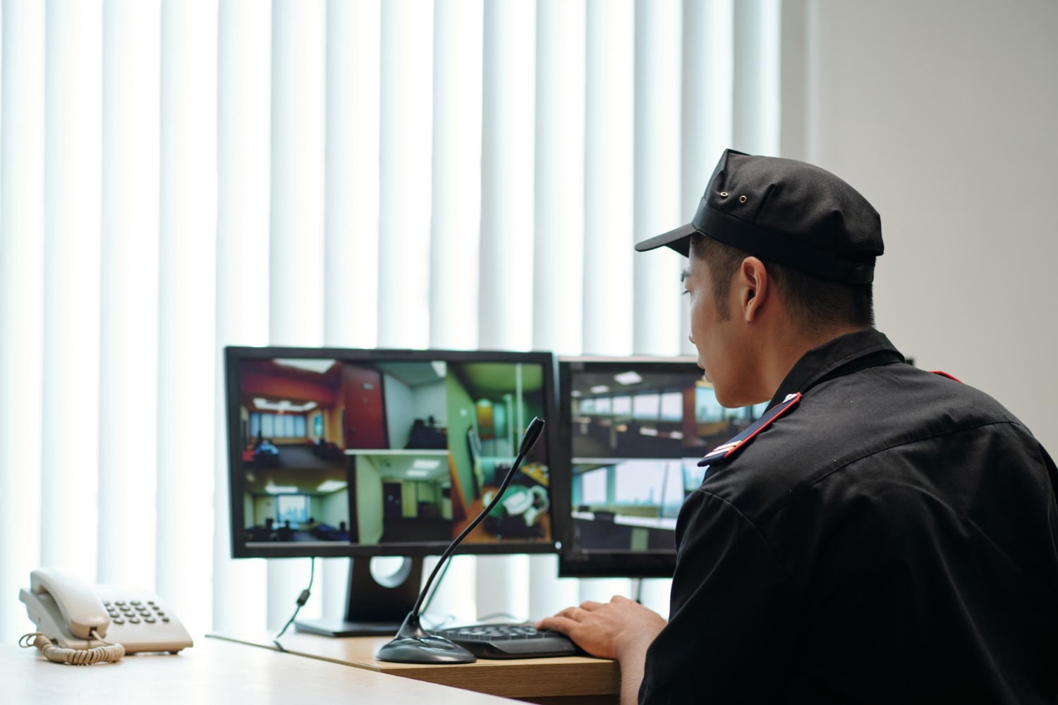 A uniformed security officer is monitoring a set of surveillance screens showing an office environment. The cloud surveillance system ensures secure office entry and workplace security through real-time video feeds.