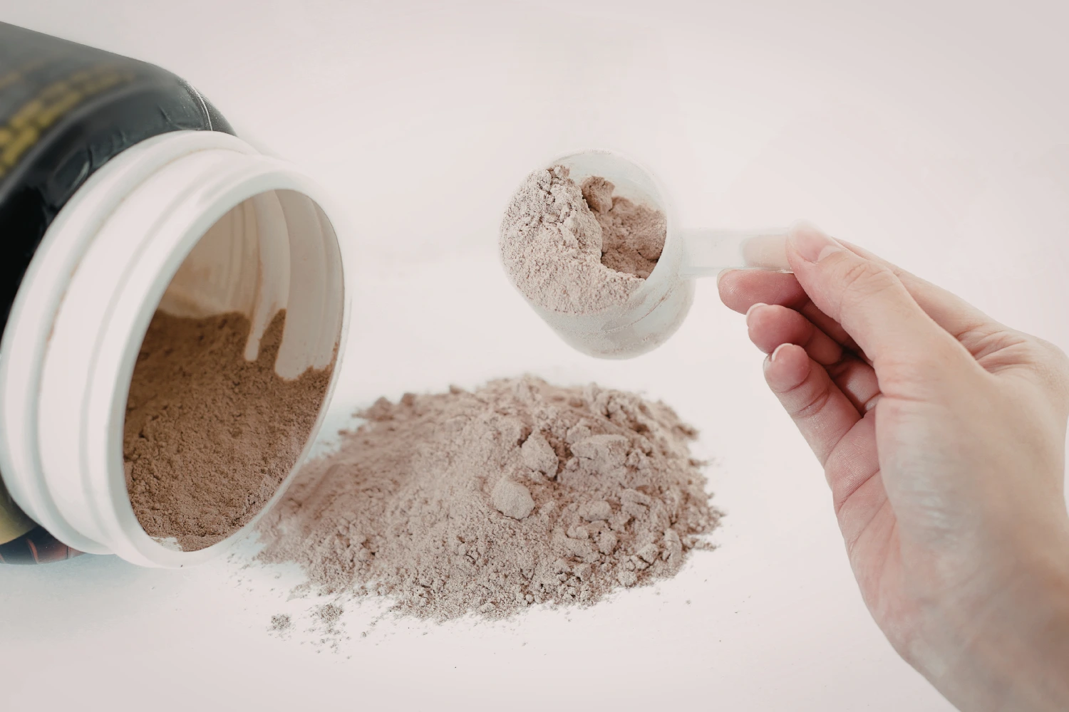 A tub of protein powder laying on its side with powder spilled out, and a hand holding a scoop of the powder.