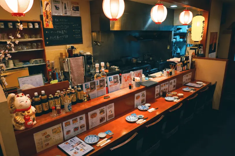 An intimate Japanese izakaya counter lined with small blue-and-white plates and chopsticks, featuring a lucky cat statue (Maneki-neko) on the left and glowing paper lanterns hanging overhead.