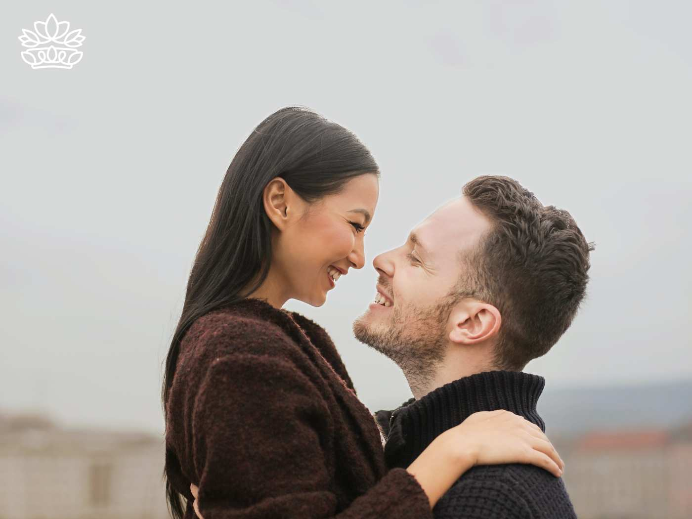 Joyful couple sharing a loving embrace outdoors, smiling at each other with genuine happiness and affection, set against a soft, muted background. Fabulous Flowers and Gifts. Delivered with Heart.