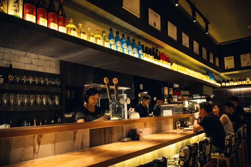 A cozy, dimly lit izakaya interior with patrons seated at a long wooden bar counter. A bartender pours a drink from a tap, while an overhead shelf displays a glowing row of various sake and liquor bottles.