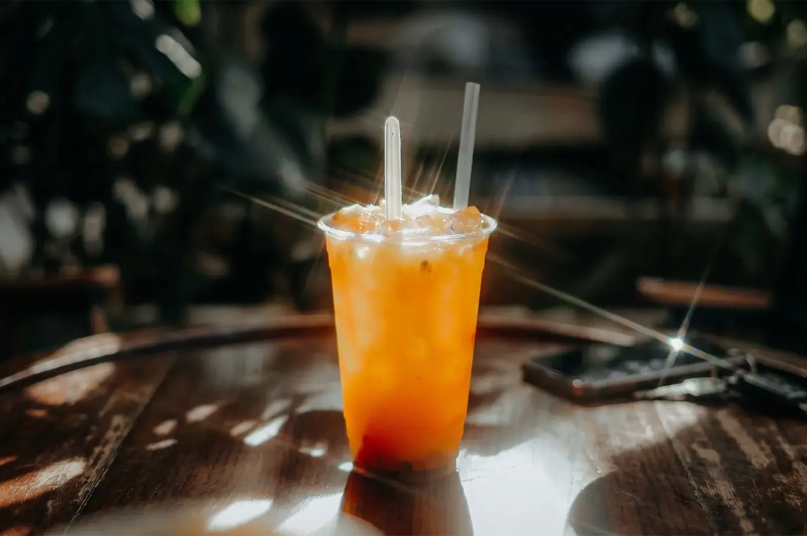 Clear plastic cup filled with an iced orange beverage and two straws resting on a dark wooden table with sun flares.