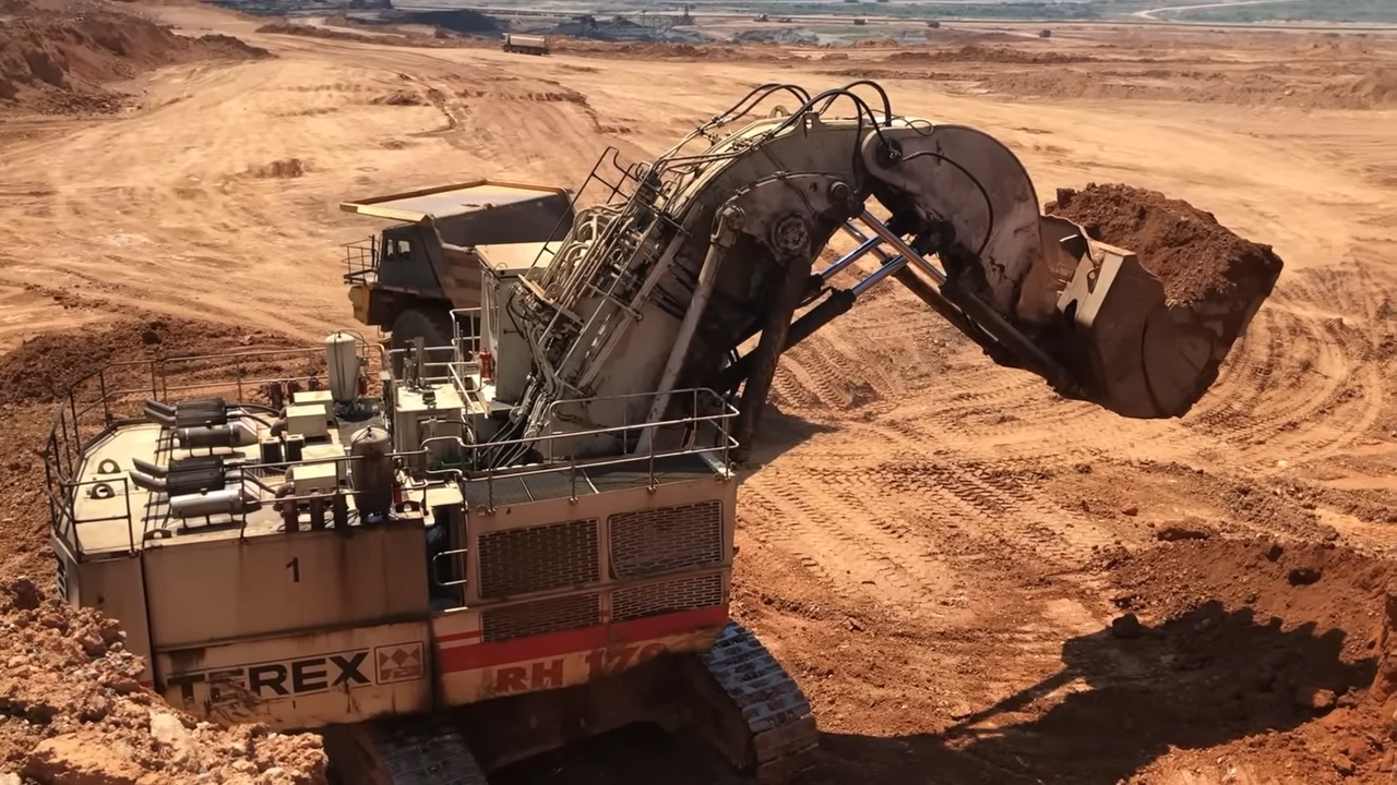 An enormous mining excavator with a full bucket of dirt operating in an open-pit mine, surrounded by a dusty, brown landscape