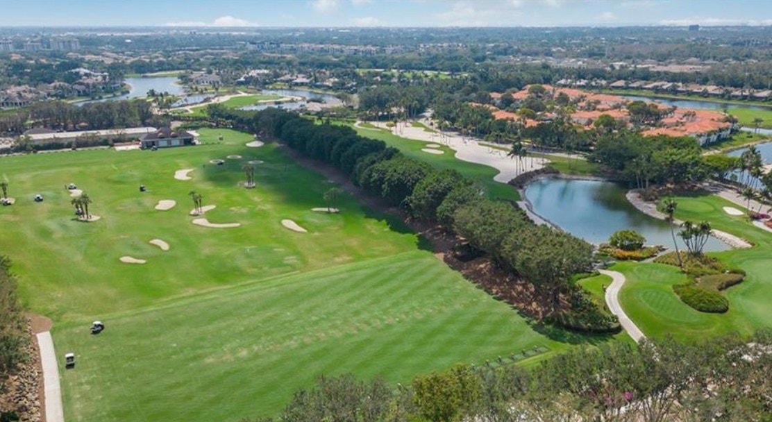 The image depicts a championship golf course at Grey Oaks Country Club, showcasing rolling greens and water hazards, with luxurious estate homes elegantly positioned in the background, embodying the essence of resort-style living in Naples, Florida. The scene captures the beauty and exclusivity of the Grey Oaks community, highlighting its exceptional lifestyle and magnificent properties.