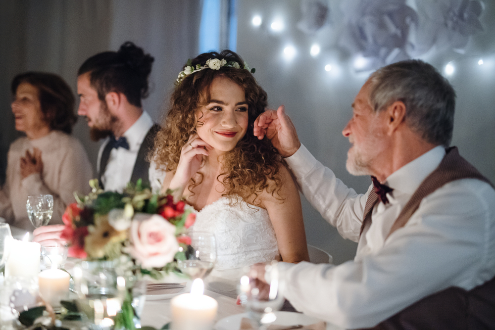 W przytulnie oświetlonym pomieszczeniu, przy elegancko udekorowanym stole weselnym, pan młody i jego żona dzielą uroczysty moment z najbliższą rodziną. Starszy mężczyzna, być może ojciec młodej, czule dotyka jej dłoni. W tle, rozmazane sylwetki gości i stoły ustawione dla reszty rodziny i przyjaciół. Choć nie widać w kadrze, okrągłe stoły są częścią starannie przygotowanego planu stołów, który uwzględnia swoich potrzeb wszystkich uczestników, w tym starsze osoby oraz tych z dalszej kolejności rodziny, a także znajomość języków obcych, aby zapewnić, że wszyscy goście na waszym weselu czują się komfortowo i są w stanie się porozumieć. Plan ten pomaga również uniknąć niezręczności siedzenia wśród obcych osób, umieszczając gości z jednej strony rodziny blisko siebie.