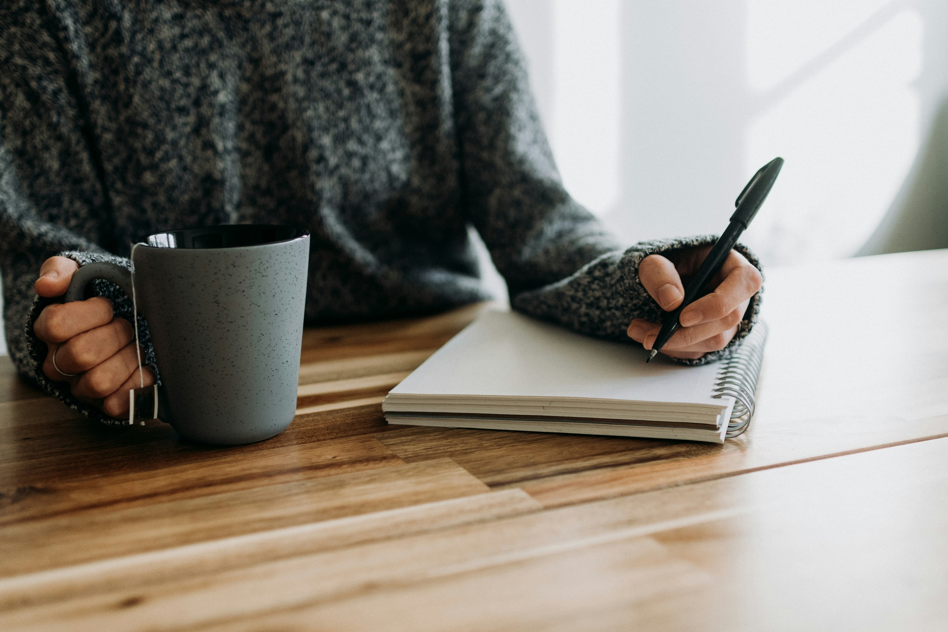 A writer sits at a desk writing in a notebook while holding a coffee cup representing us writing contests.