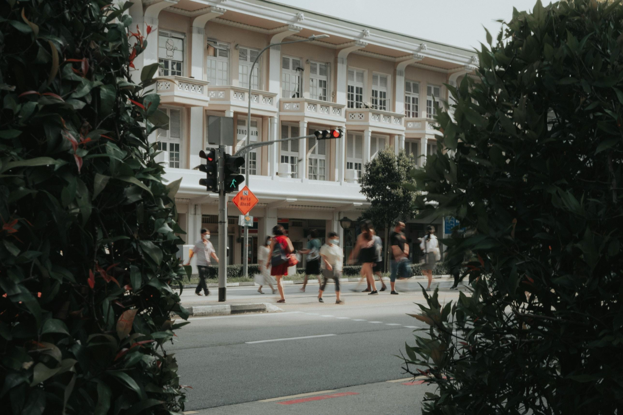 Framed by dark green foliage in the foreground, a group of pedestrians crosses a city street at an intersection marked by traffic lights and signage. The backdrop features a grand, cream-colored building with classical architecture, shuttered windows, and balconies, adding a historic feel to the busy urban scene.