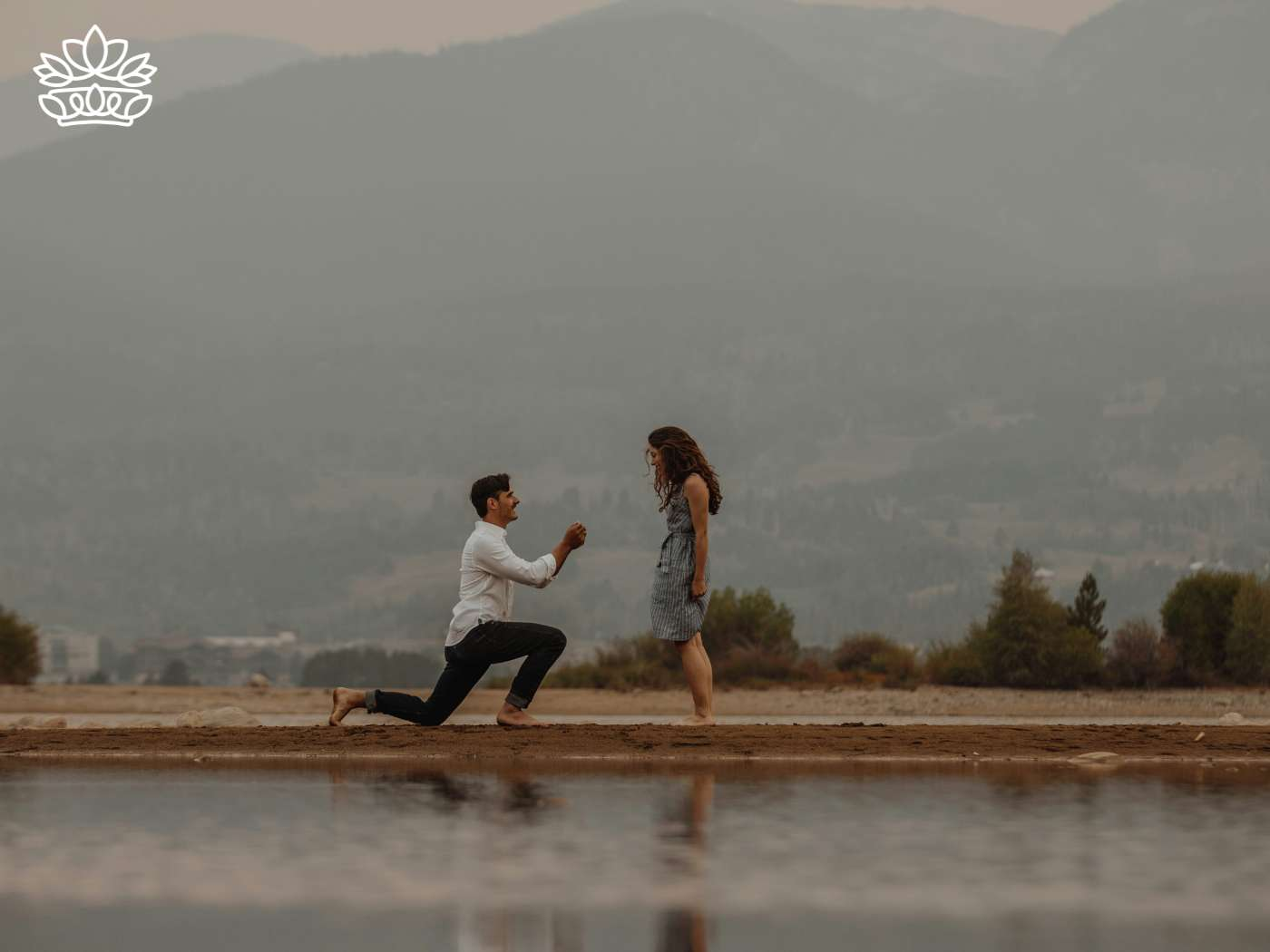 A happy couple during a heartfelt proposal, with the man on one knee by a serene lakeside, a backdrop of misty mountains enhancing the moment. This scene captures the essence of the best engagement gift, offered in a sweet way from the Engagement Collection at Fabulous Flowers and Gifts