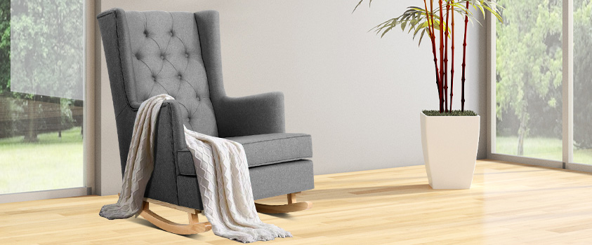 A grey rocking fabric recliner, with a beige knitted blanket draped over one arm, set in a bright living room with white walls, light floors, two large windows and a pot plant.