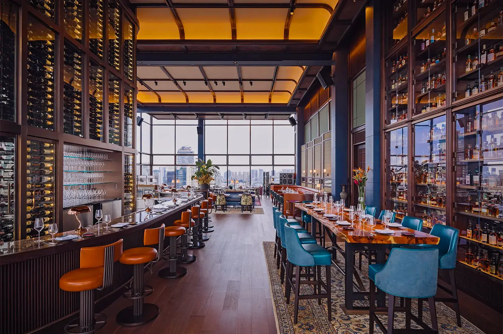 Luxurious rooftop bar with warm lighting, a long counter with orange stools on the left, teal chairs around tables on the right, and city views through large windows.