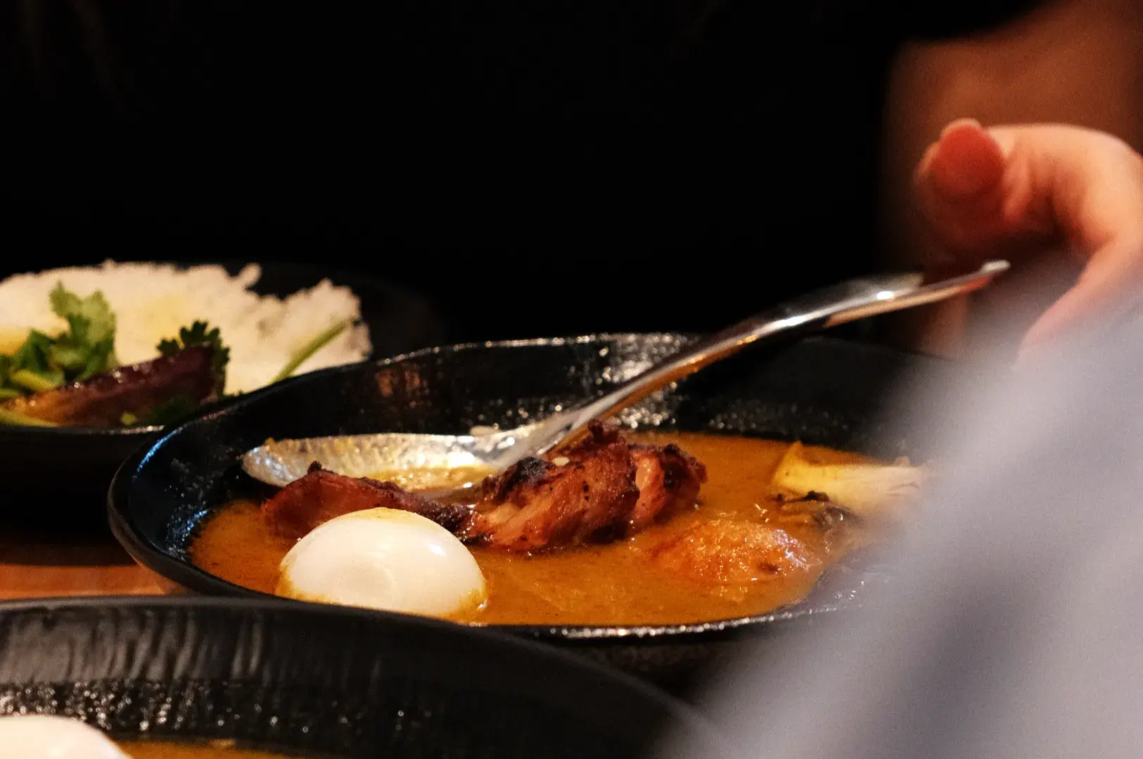 A close-up of a bowl of curry with chicken, boiled eggs, and a spoon, alongside a plate of rice and greens.