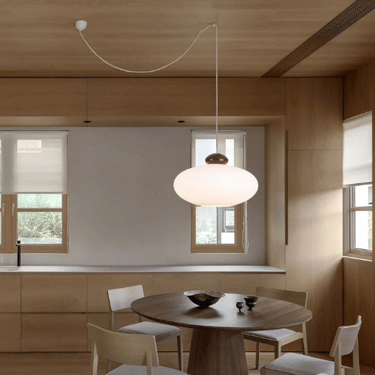 Minimalist dining area with a round wooden table, four chairs, and a large frosted glass pendant light hanging above, set against light wood cabinetry and windows with roller blinds.