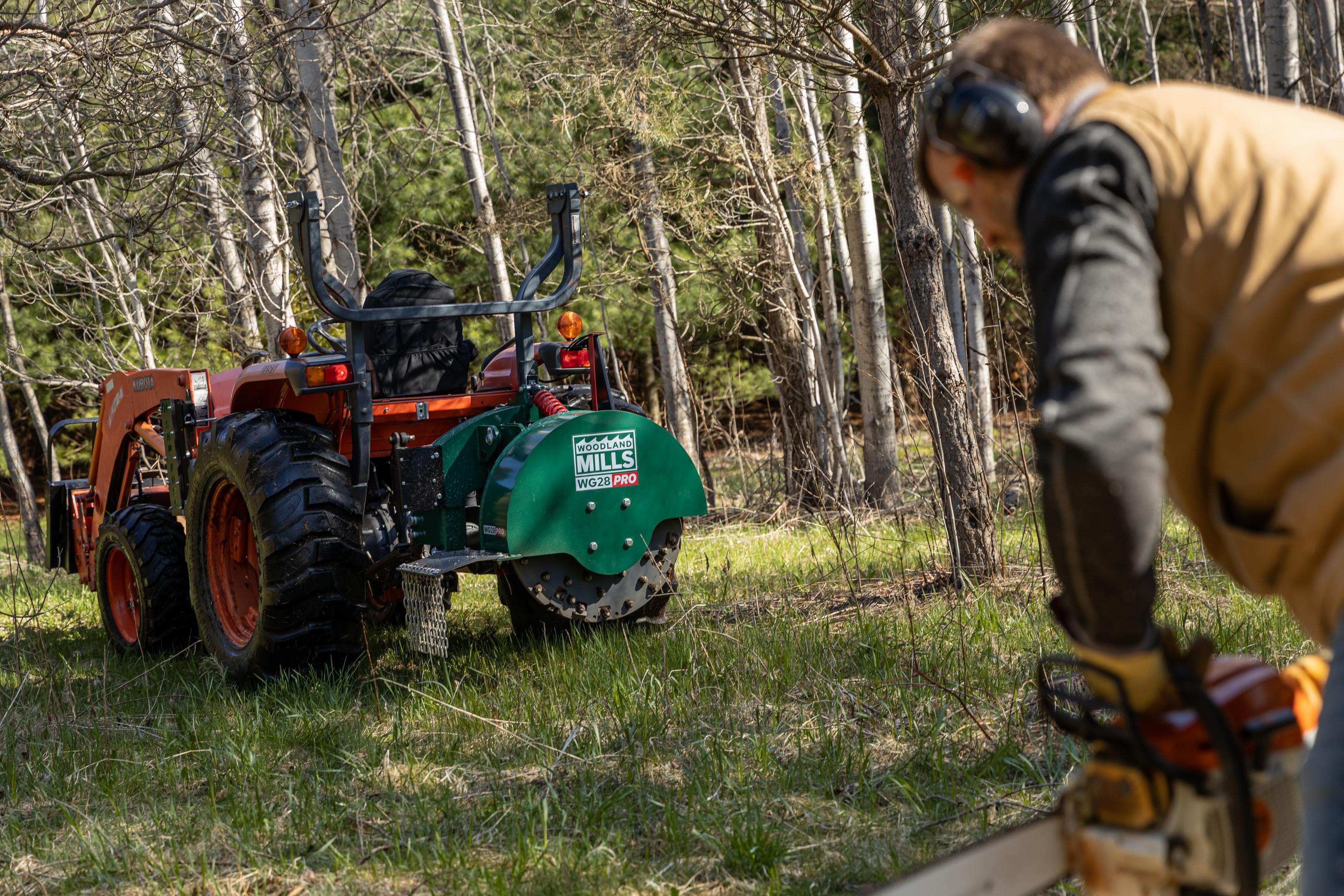 A Woodland Mills WG28 PRO Stump Grinder attached to the back of an orange tractor. 