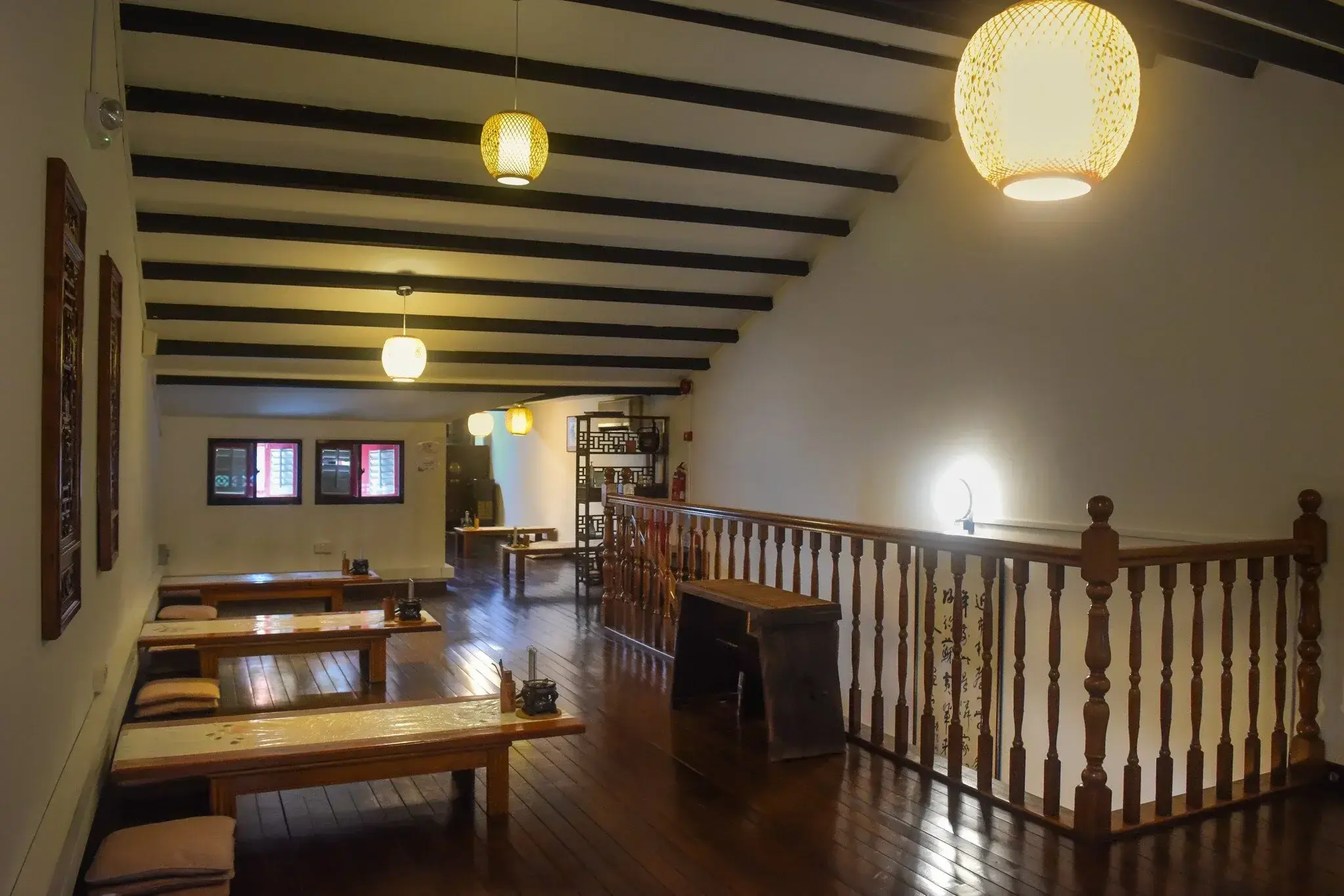This interior space features a warm, traditional aesthetic with low wooden tables, floor seating, and dark timber ceiling beams. Softly glowing globe lanterns hang from the slanted ceiling, illuminating the polished wood floors and a classic balustrade.