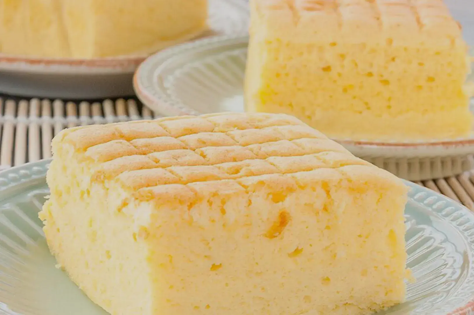 A close-up of a slice of fluffy, Japanese cotton cheesecake on a plate, with additional slices in the background. The cheesecake has a light, airy texture and a golden top with a crisscross pattern.