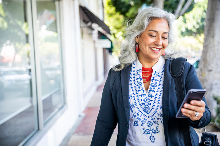 Woman with long gray hair and a red necklace smiling at her cell phone.