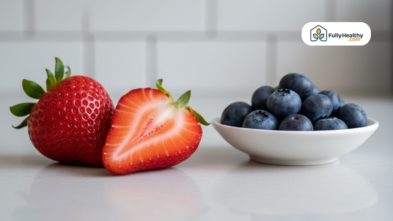 Whole and sliced strawberry beside bowl of blueberries on clean kitchen counter