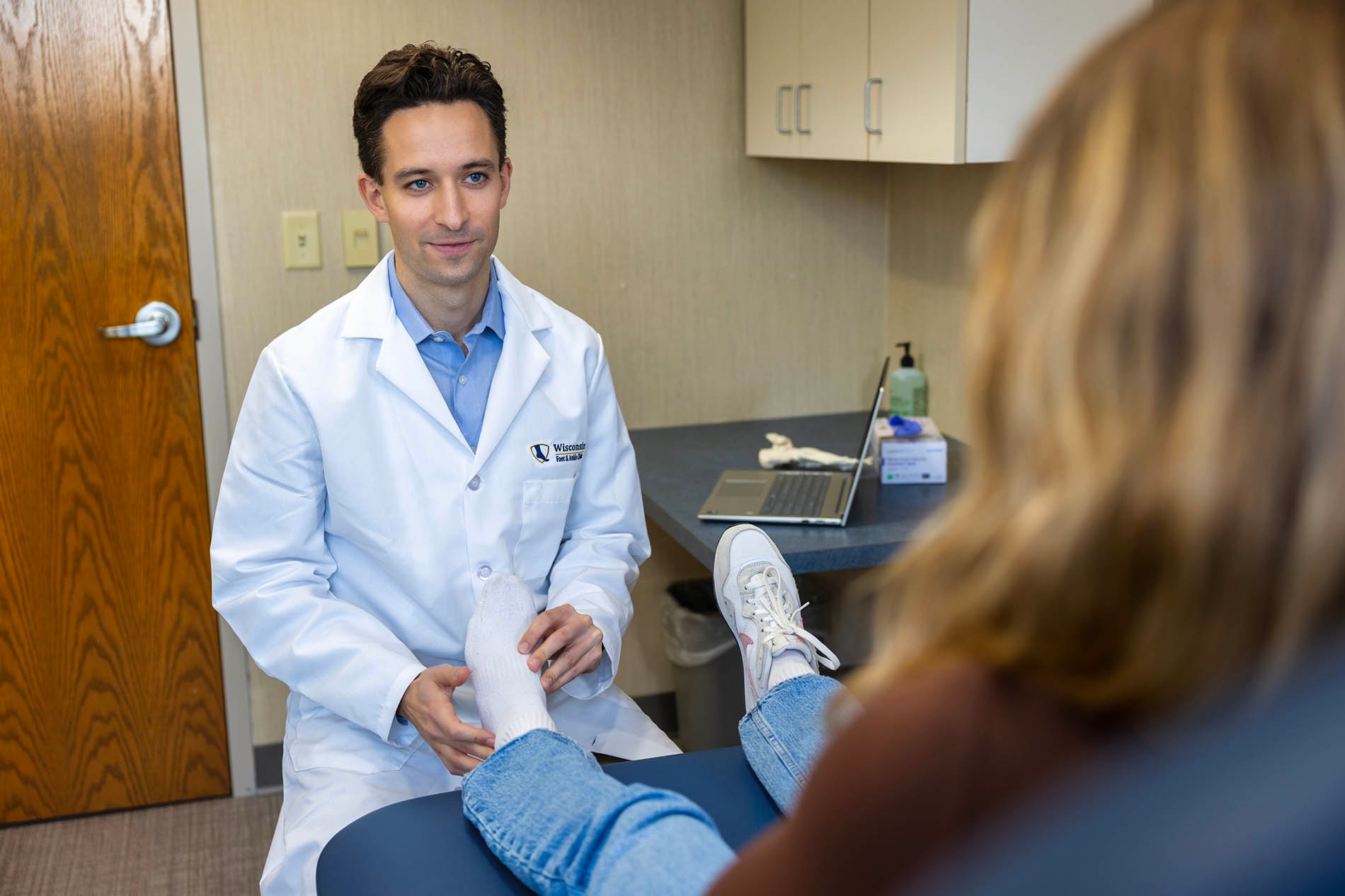 Foot and ankle doctor examines a patient’s foot on an exam table in a medical office, with a laptop and supplies on the counter nearby.