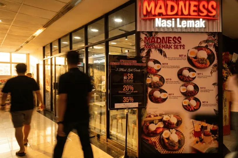 The exterior of the Madness Nasi Lemak restaurant in Fortune Centre, featuring a bright red and white neon sign. A large, colorful menu board displays photos of various Nasi Lemak meal sets alongside a standing sign advertising opening promotions, while blurred pedestrians walk past the glass storefront.