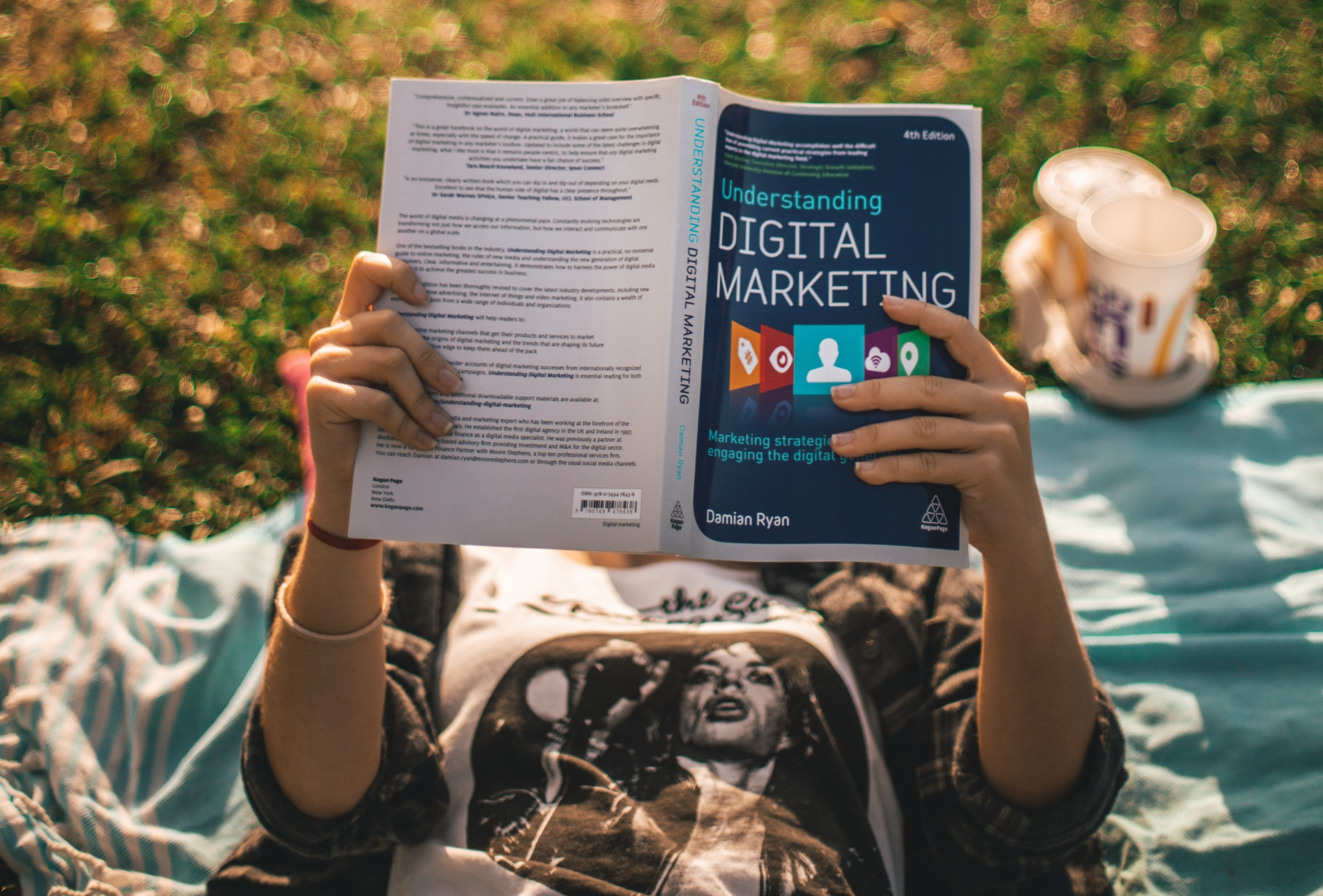 Une jeune femme, allongée sur du gazon, lit un livre sur le marketing digitale. Le marché de la publicité classique et digitale sont toujours en plein expansion. La publicité digitale est importante pour la promotion des marques