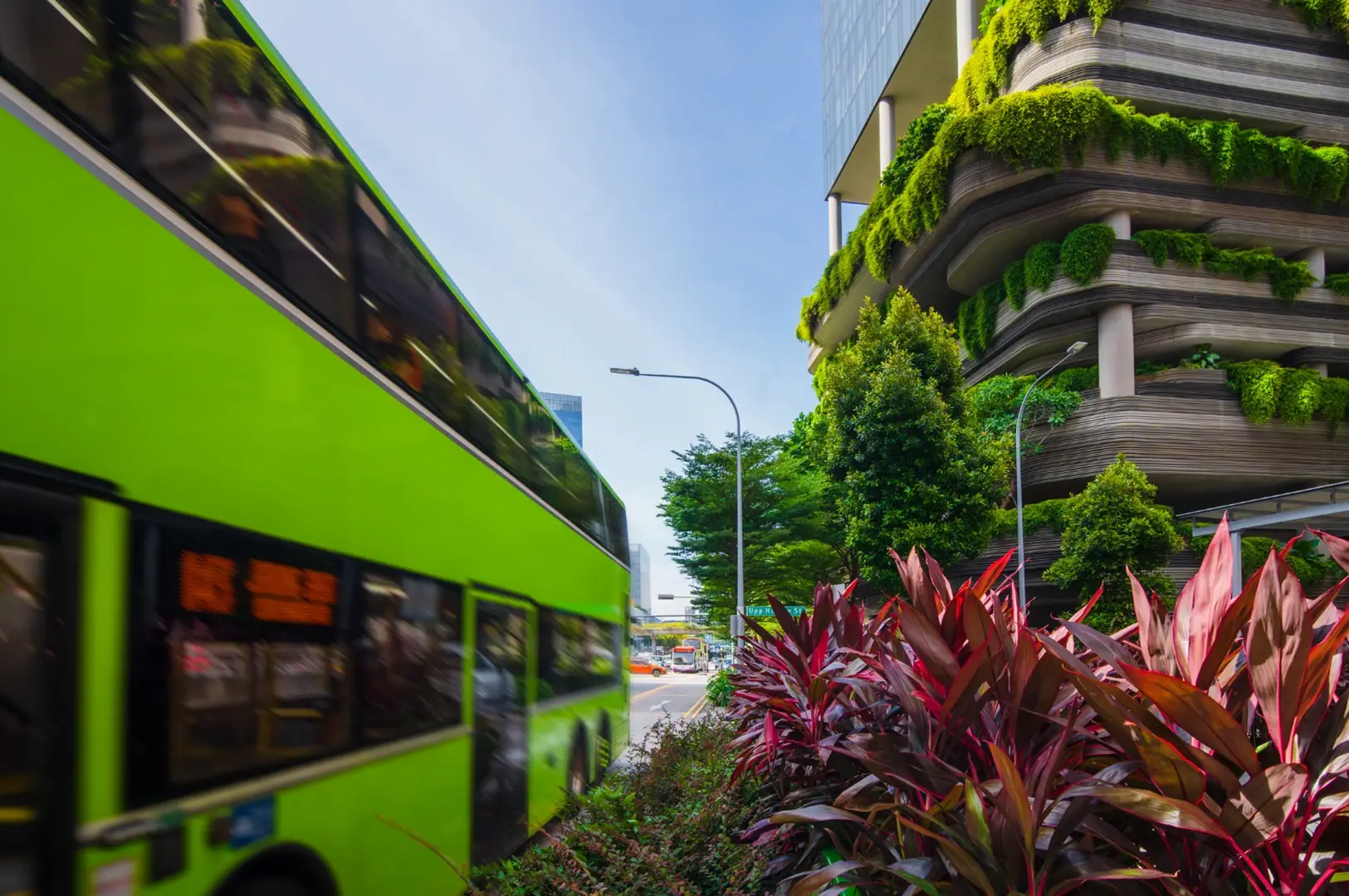 A bright green double-decker bus drives past a modern building with cascading greenery and red foliage in the foreground.