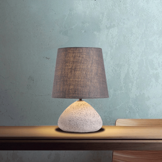 Modern farmhouse table lamp with a textured stone base and a dark fabric shade, placed on a wooden table against a rustic light green wall, creating a warm and cozy ambiance.