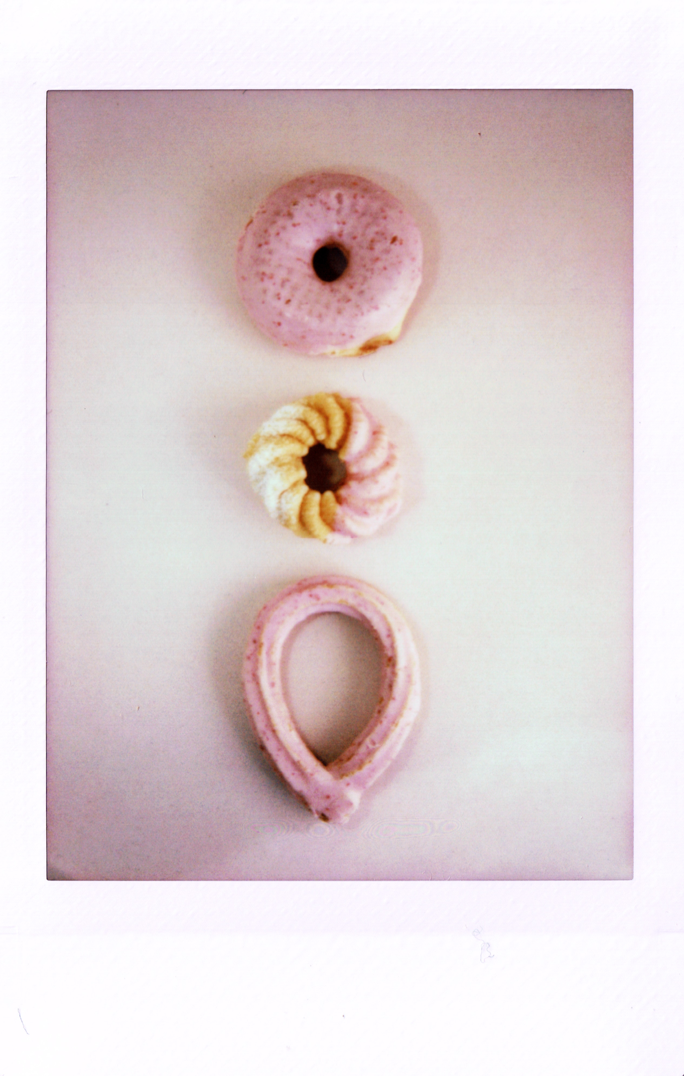 A picture of three doughnuts with pink icing, the top one is a normal ring doughnut, the middle is a french doughtnut, and the bottom is a looped chouro. 