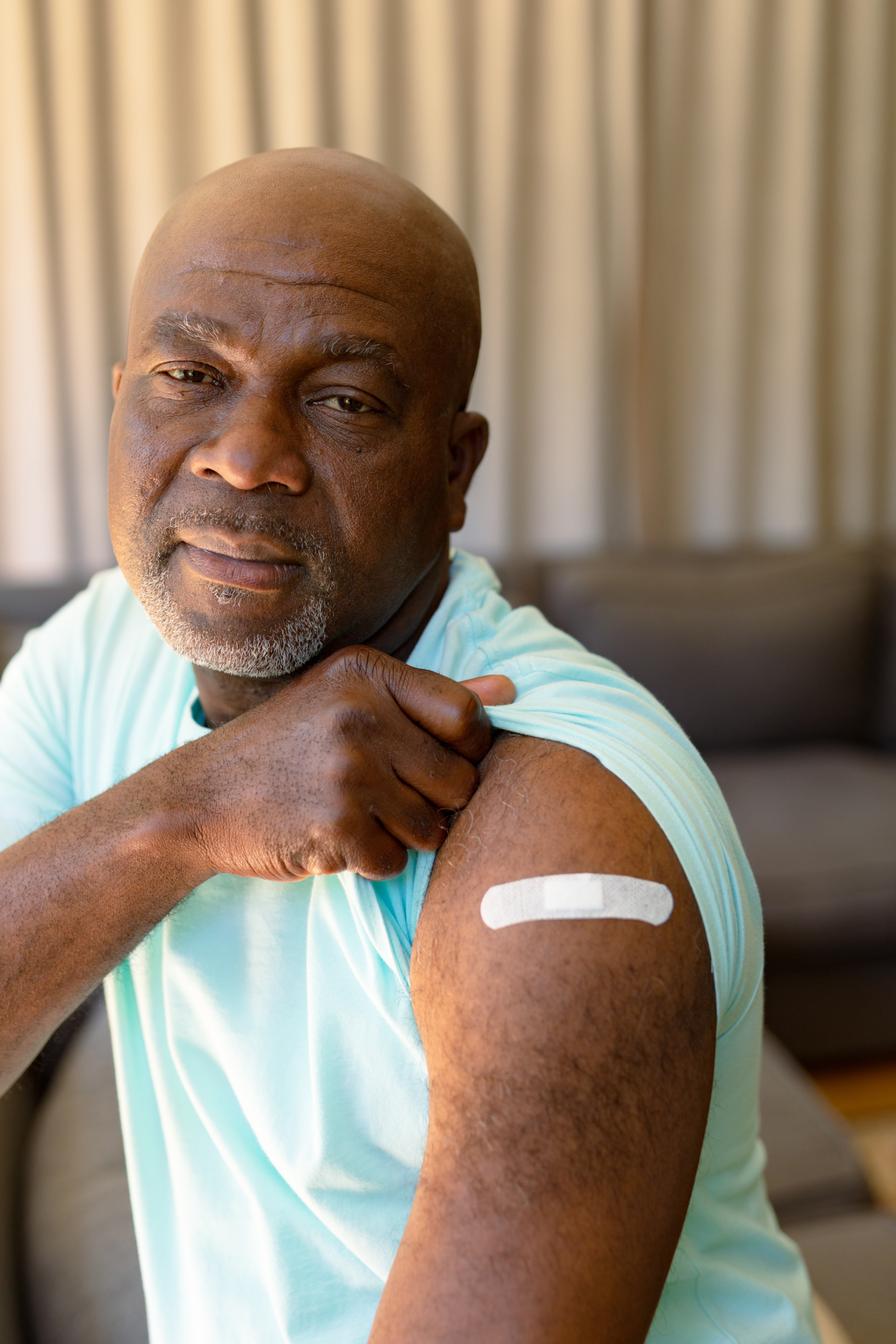 Image shows a balding, black man in his 50s. He's wearing a light aqua-coloured t-shirt. He is holding up one sleeve to show a plaster on his upper arm.