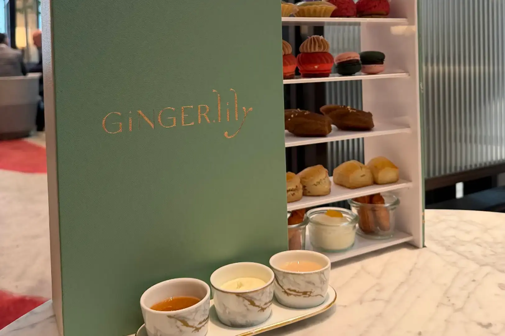 A close-up view highlights a sage-green book-style afternoon tea set with "GINGERLILY" embossed in copper foil across its cover. The open side reveals stacked white shelves displaying delicate pastries, macarons, baked scones, and small glass dessert cups, with three marble-patterned condiment pots arranged on a tray in the foreground.