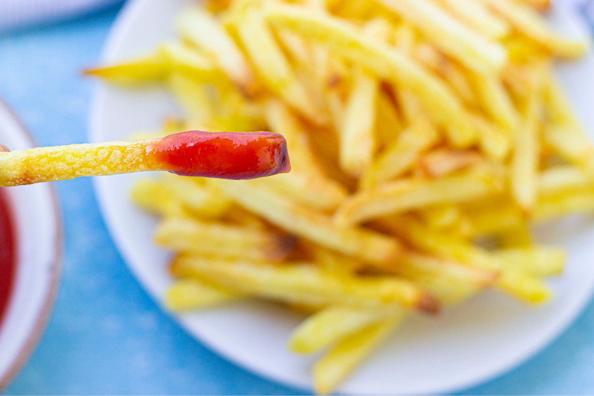 A piece of fries with ketchup and a plate full of fries at the back.