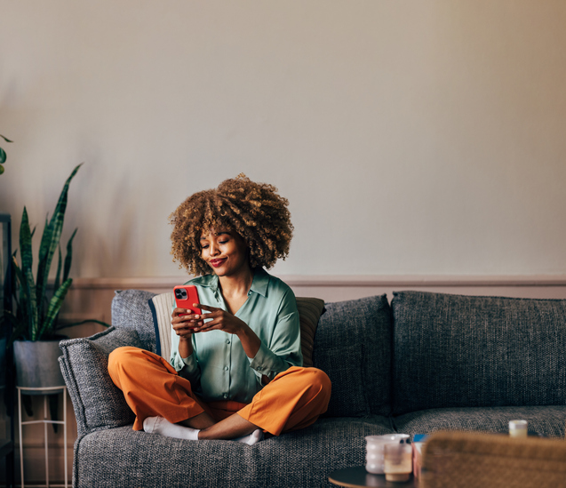 Happy young woman with lush curly hair relaxing on her sofa with her smartphone.  