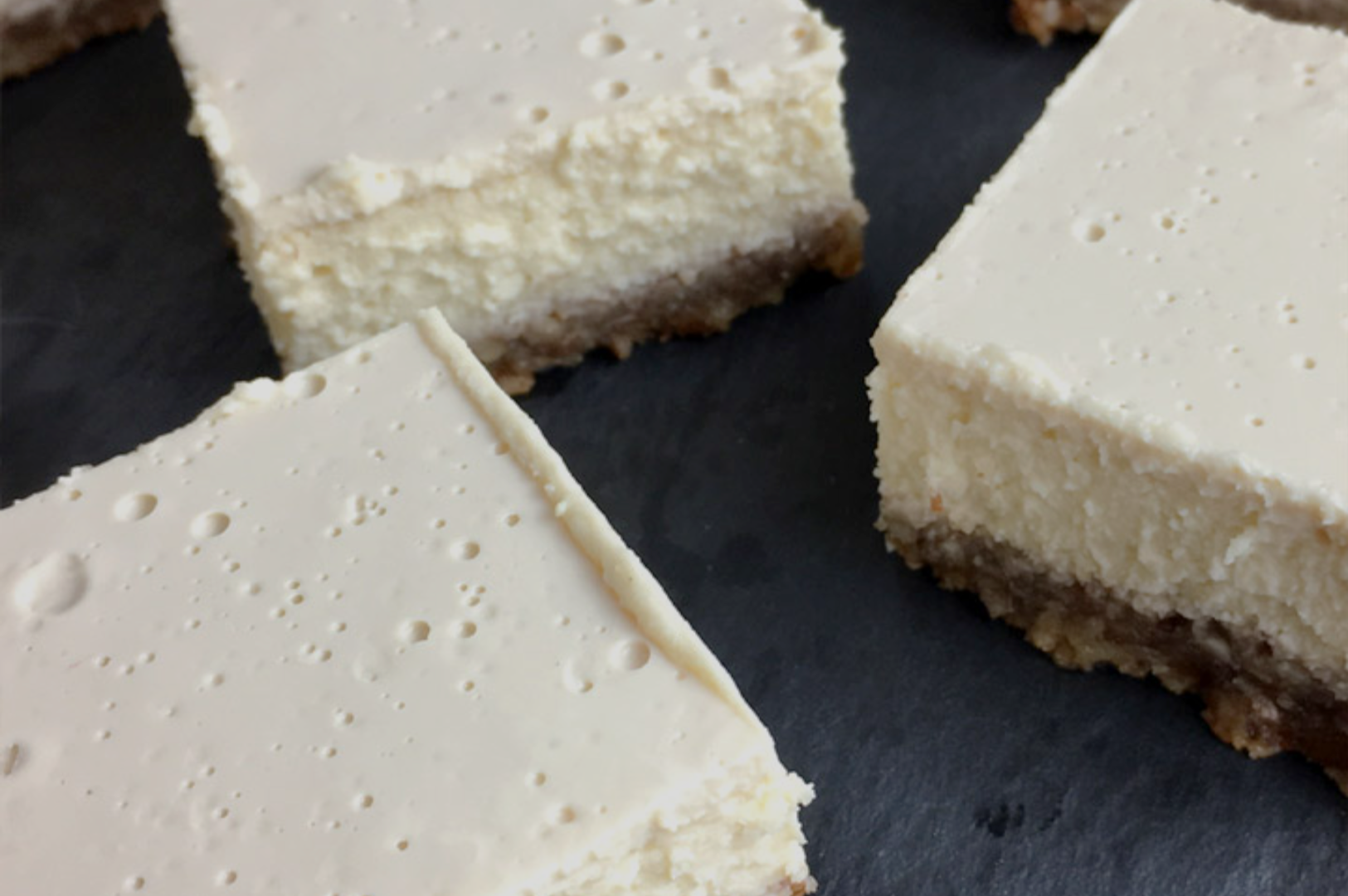 Four creamy cheesecake squares with a light, bubbly surface and a brown crumbly base, arranged on a dark slate background.