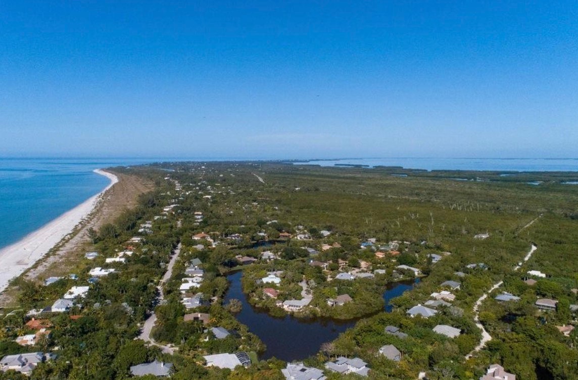 The image depicts a luxurious beachfront home with elevated construction, surrounded by lush tropical landscaping and offering stunning Gulf views. This exceptional near beach estate is located in Sanibel's Gulf Pines neighborhood, featuring deeded beach access and a private beach path for residents.