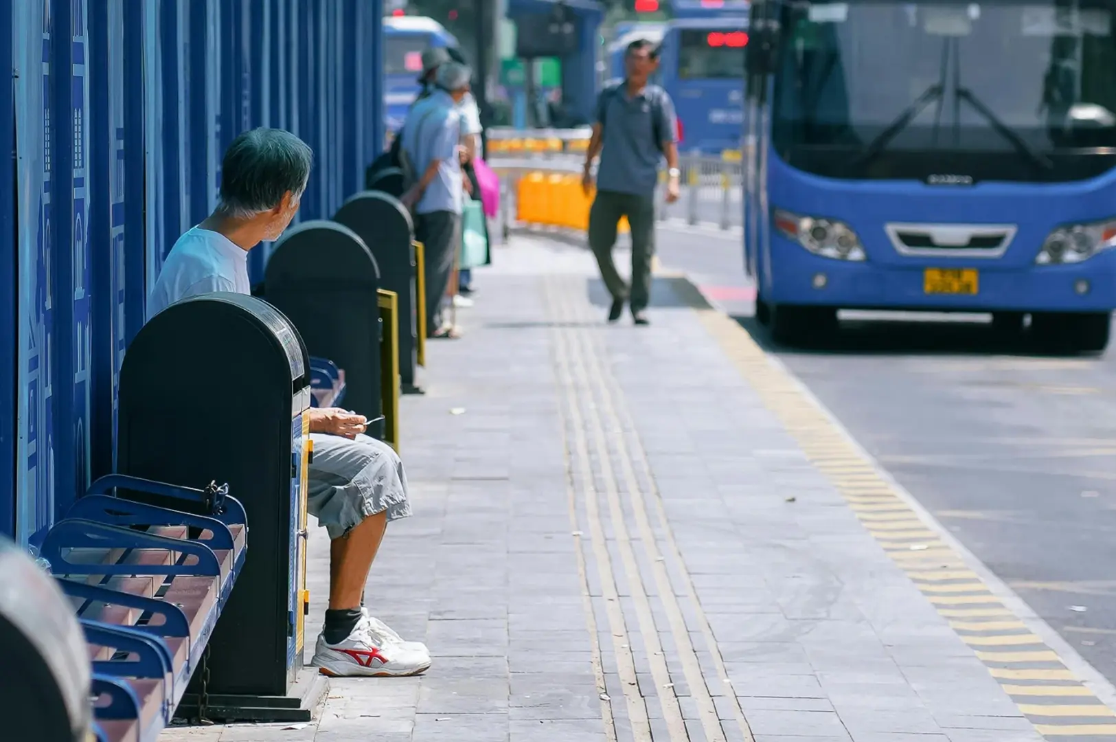A blue bus stop shelter with route numbers displayed overhead. People are waiting on the sidewalk next to a blue bus.