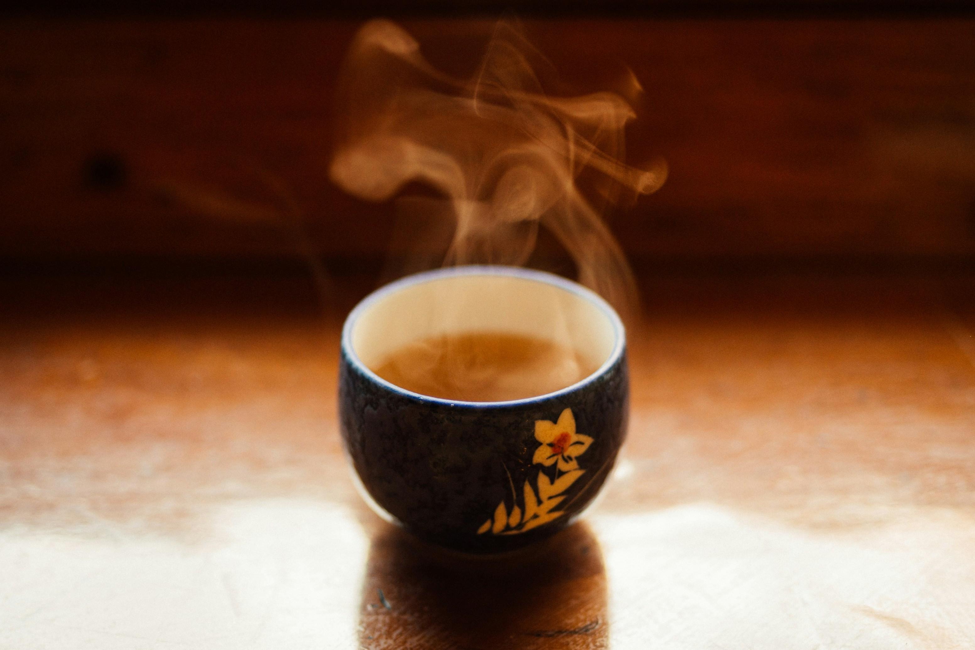 A small, ornate ceramic cup sits on a wooden surface, filled with a warm liquid that releases soft plumes of steam. The dark blue vessel is decorated with a delicate yellow flower, all captured in a warm, shallow-focus glow.