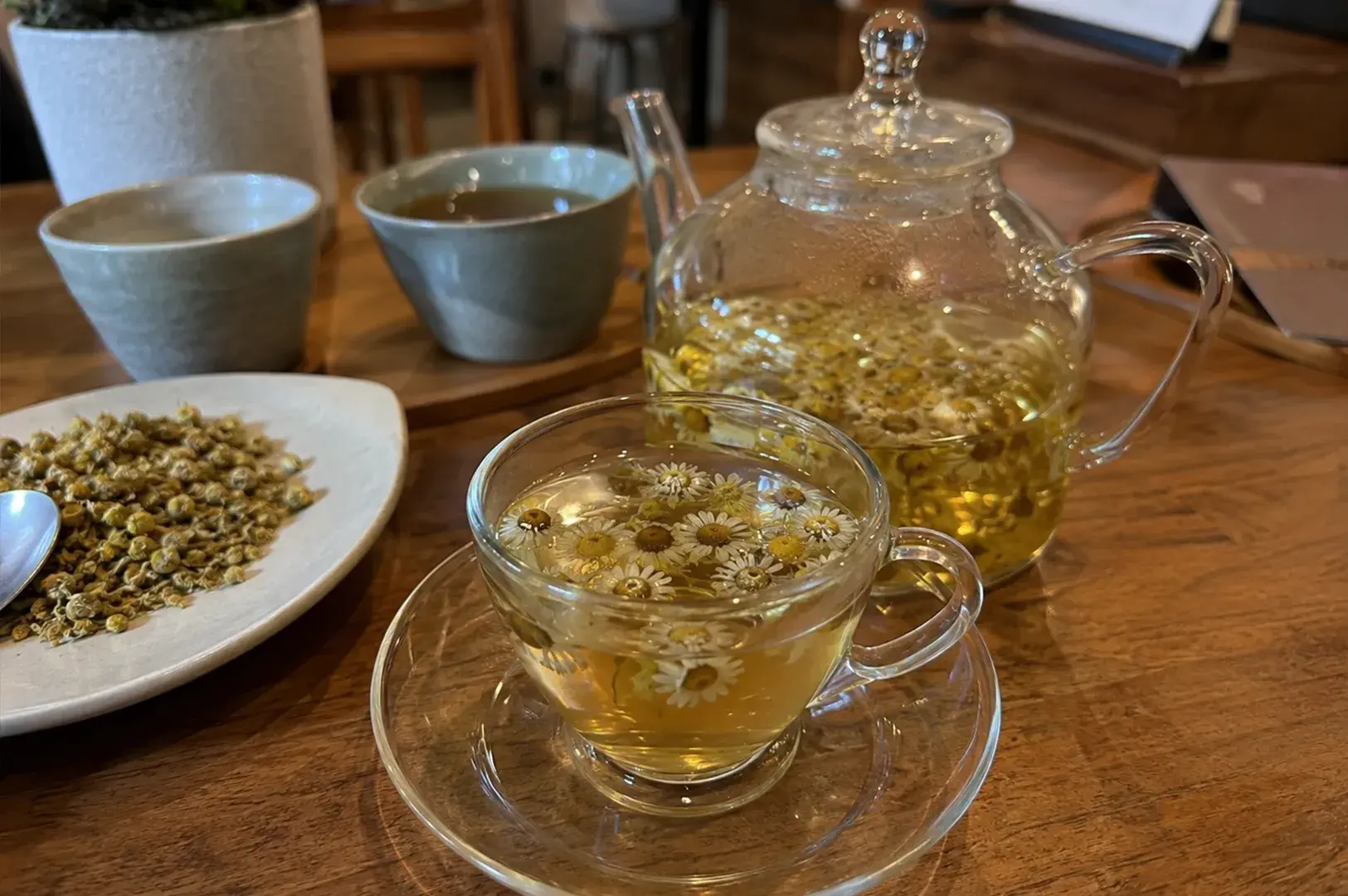 A close-up, eye-level shot captures a cozy tea setting on a warm wooden table, featuring a clear glass teacup filled with light golden chamomile tea and floating whole chamomile blossoms in the foreground. Behind it sits a matching clear glass teapot infusing more loose-leaf tea, alongside two ceramic cups—one grey and one light blue—and a shallow white plate piled with dried chamomile flower buds to the left. The background is softly blurred, hinting at an inviting, warm cafe ambiance.
