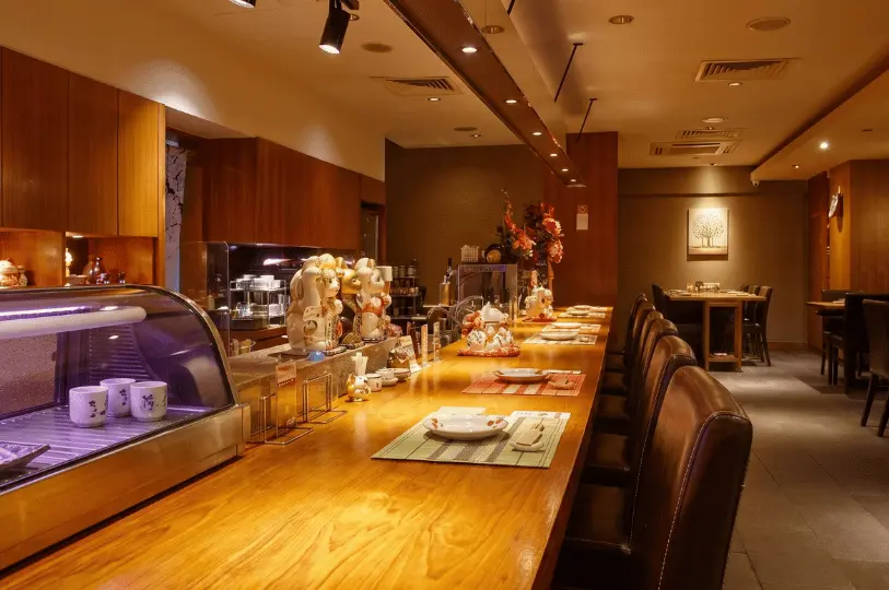 The interior of a cozy Japanese restaurant featuring a long, polished wooden counter lined with leather chairs and a playful collection of maneki-neko lucky cat figurines on display.
