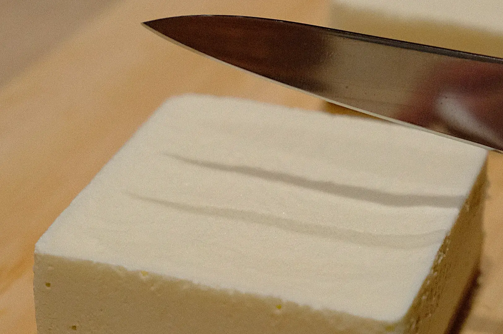 A close-up of a knife about to slice a block of tofu on a wooden cutting board.