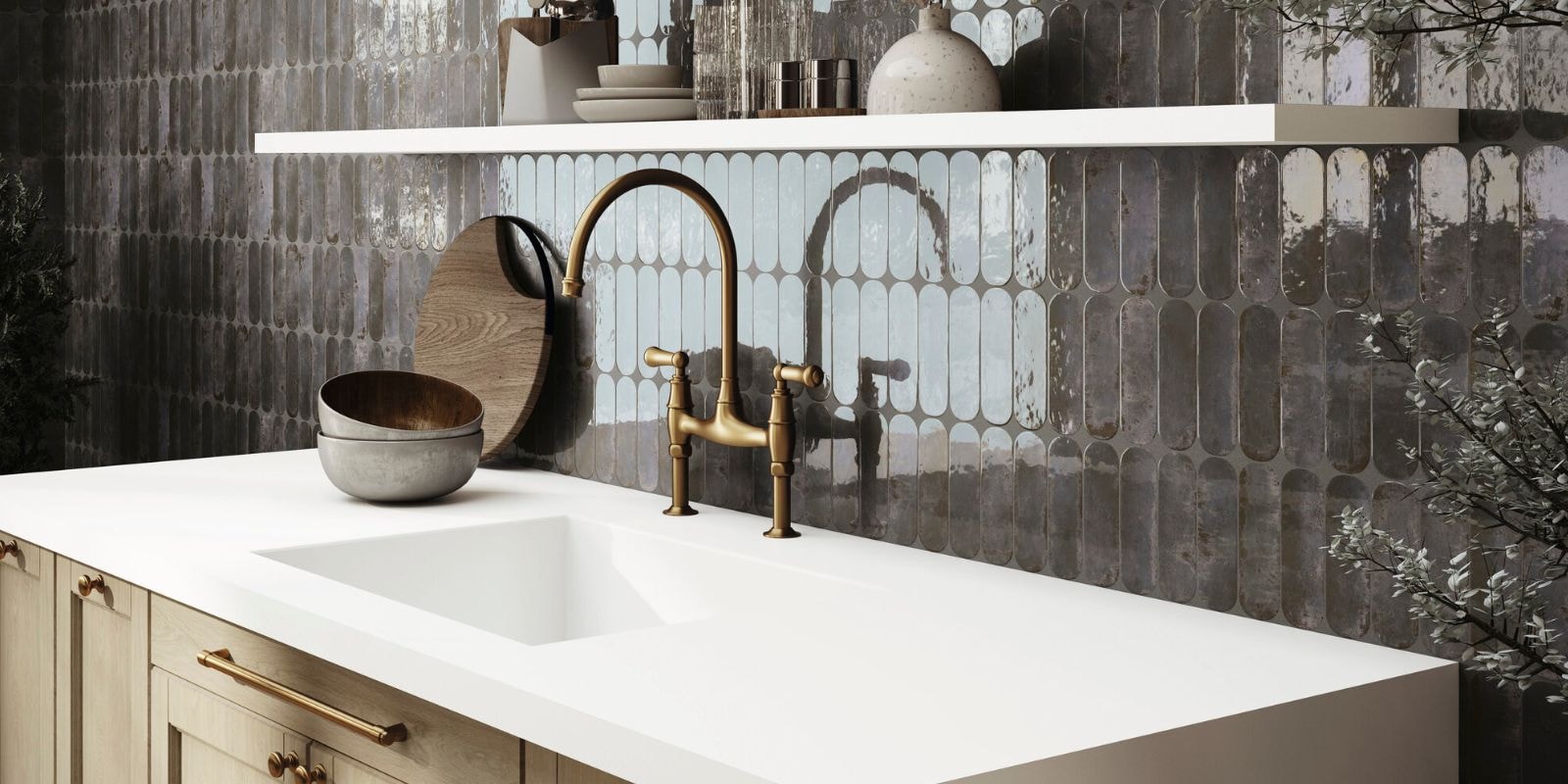 Glossy elongated oval and rectangular backsplash tiles in shades of grey and blue behind a white countertop and brass faucet.