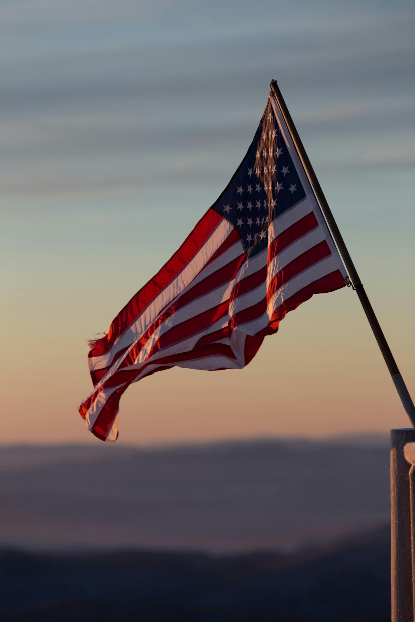 American flag waving on a pole at sunset, silhouetted against a sky with soft gradient hues.