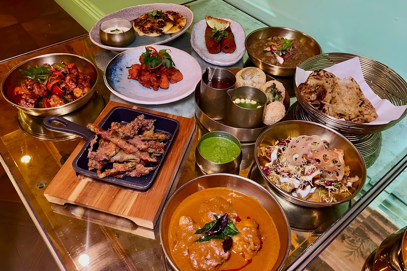 An array of Indian dishes on a table includes curry, skewers, breads, mixed starters, and dips. The setting is vibrant and appetizing.