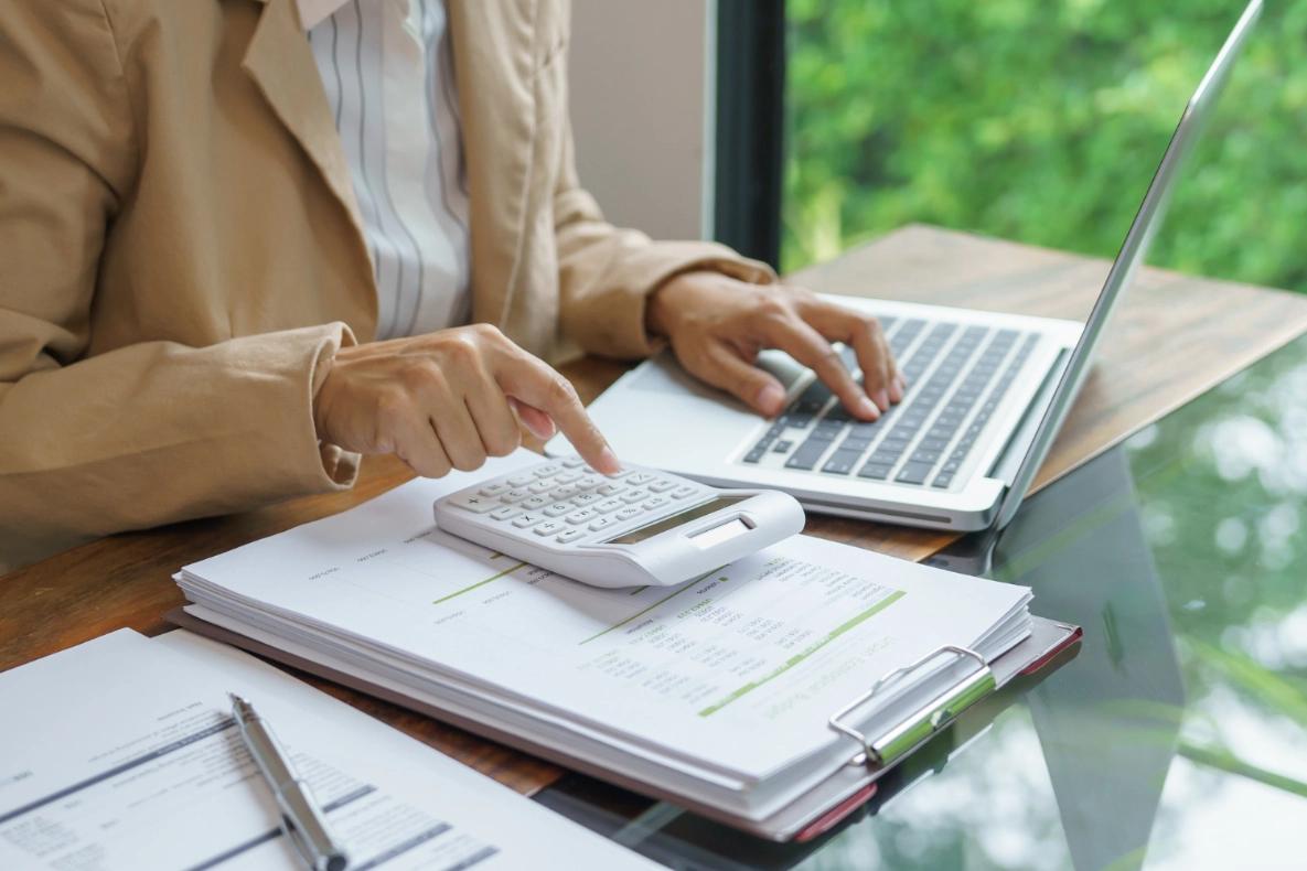 A person in a tan blazer uses a calculator and types on a laptop while reviewing documents on a desk with a blurred green background.