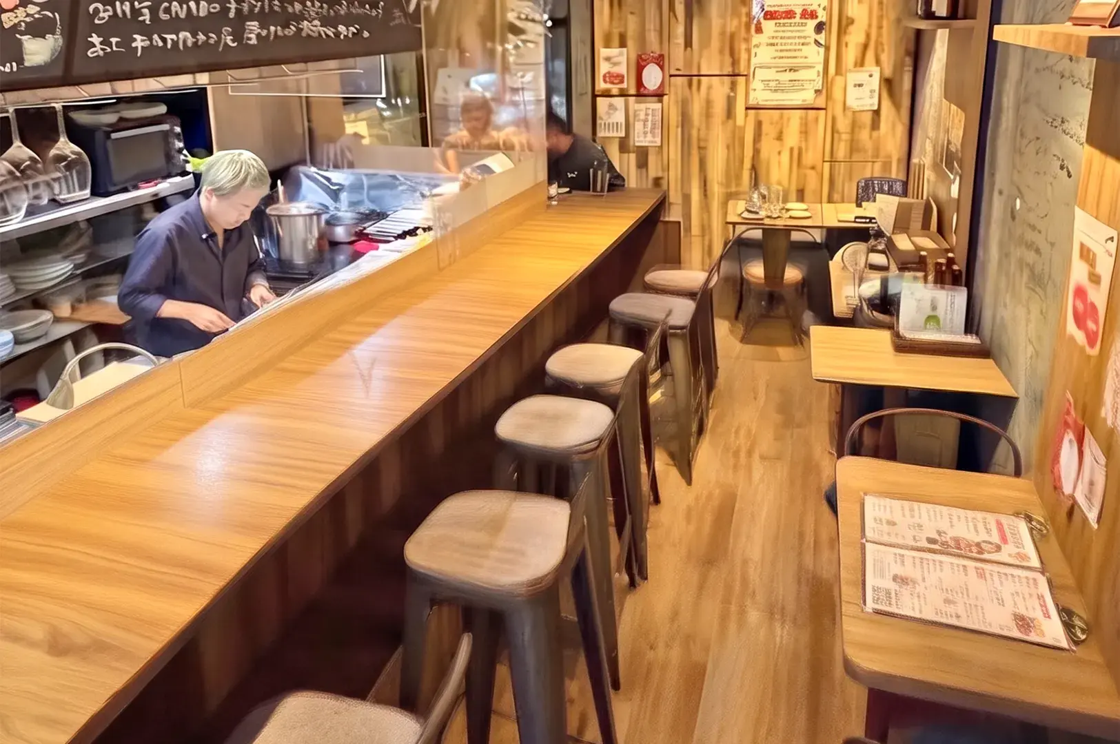 Interior of Al Solito, a cozy Japanese restaurant featuring a wooden counter bar, stools, and a chef preparing food.