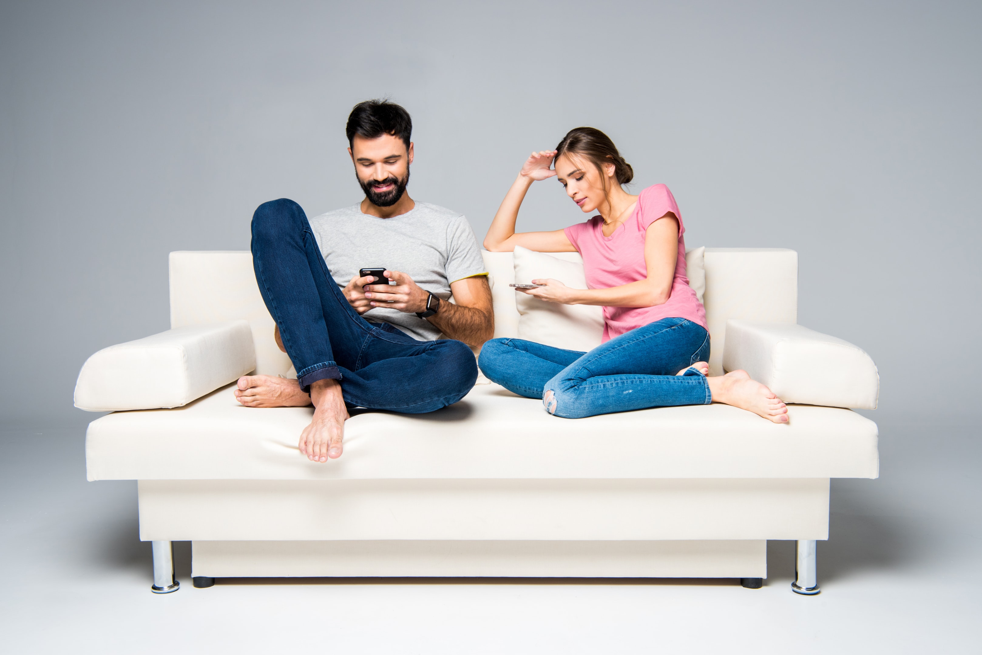 A well-dressed professional couple sitting on opposite ends of a modern couch, both absorbed in their devices despite being in the same space.